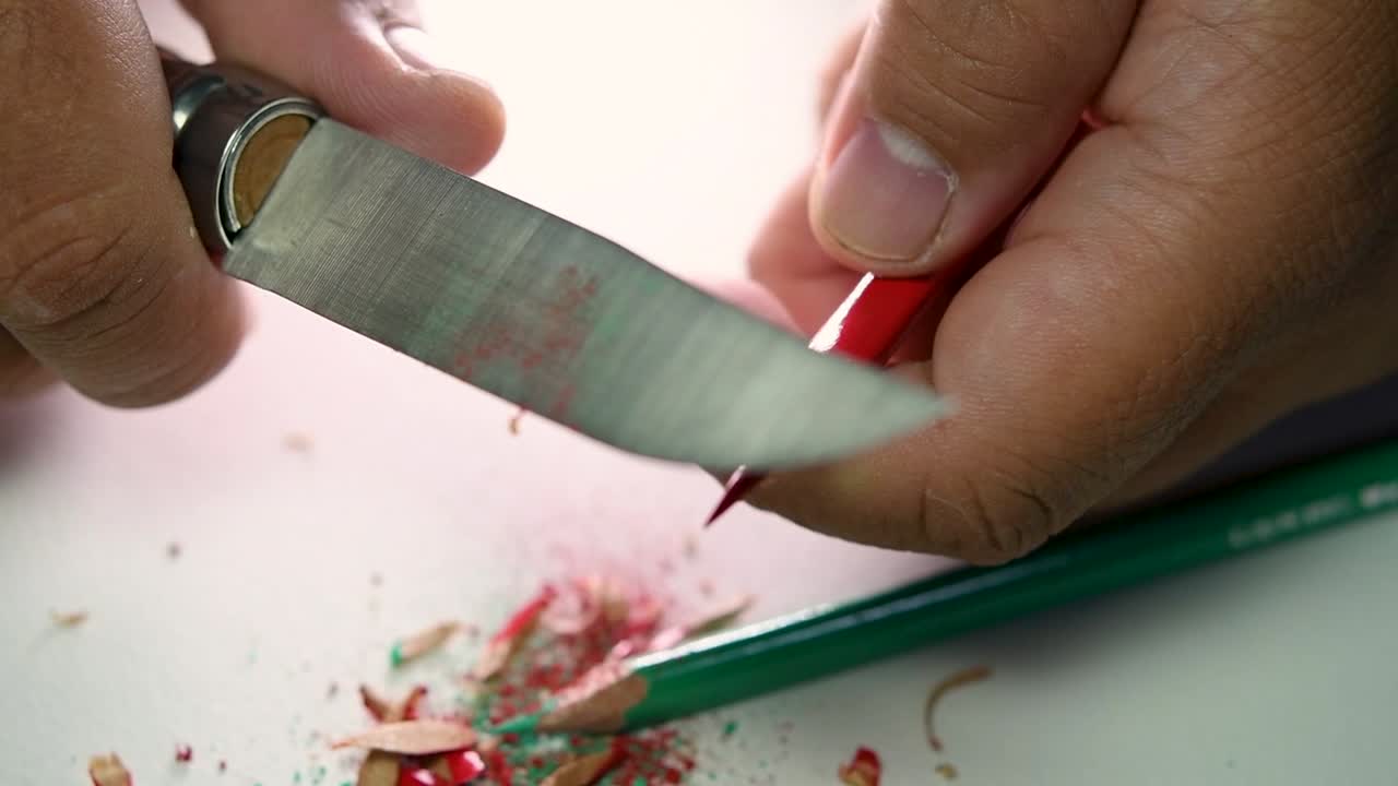 Footage of hands slowly sharpening a pencil and some coloured pencils with a sharp knife