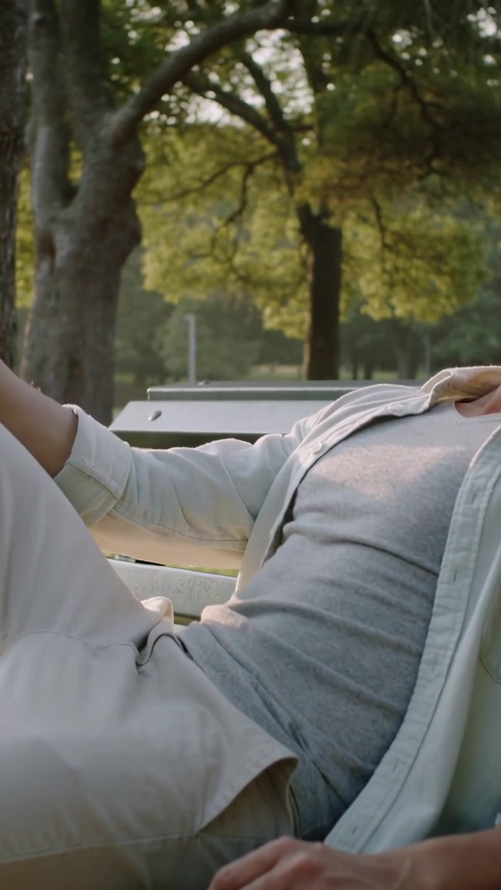 Vertical video: Reclining man wearing light top settling on park bench as sun filters through trees