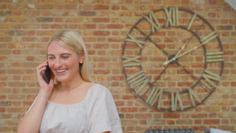 Young-Woman-Standing-By-Brick-Wall-With-Clock-At-Home-Talking-With-Friends-On-Mobile-Phone