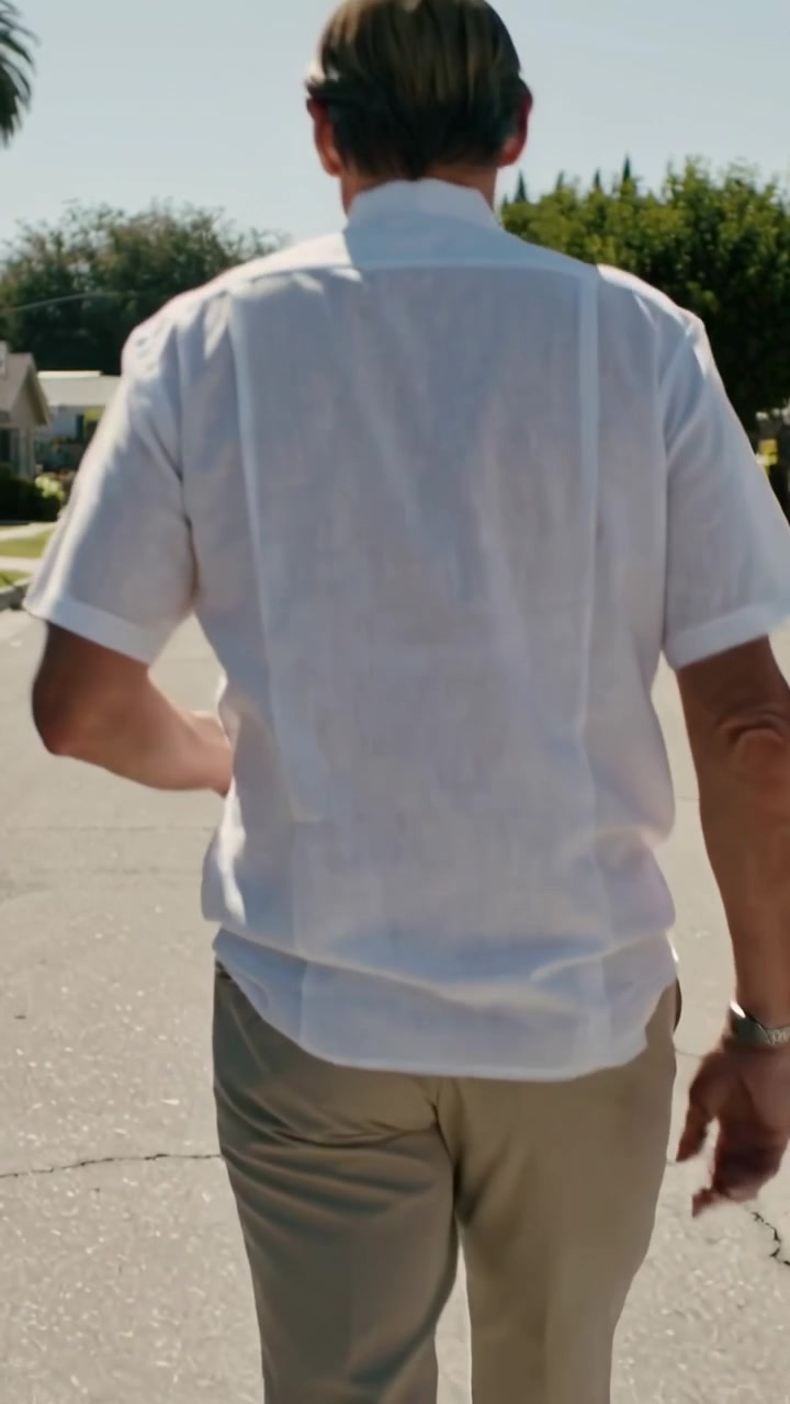 縦型動画：サングラスをかけた男性がヤシの木が並ぶ郊外の通りを歩き始める様子を捉えたカメラ。
