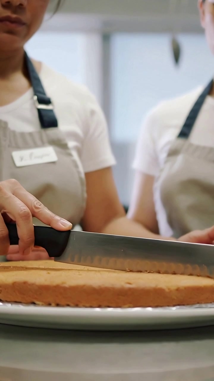 Vertical video: Placing serrated knife, two bakers leveling cake for even top at counter in aprons