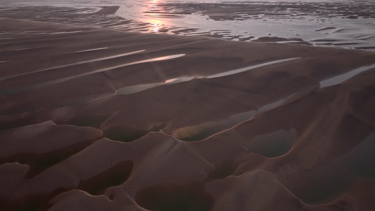 Sunset reflecting off long ridged sand patterns and pools at low tide. Reverse flight over.