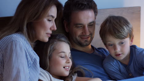 Close-up-view-of-caucasian-parents-sitting-on-the-bed-with-their-little-cute-son-and-daughter,-watching-something-on-the-tablet-and-talking-at-night