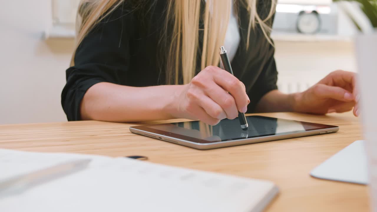 mujeres de negocios que trabajan en la tableta en la oficina. atractiva mujer rubia con cabello largo en traje elegante sentada en un escritorio de madera con computadora y concentrándose en el trabajo en la tablet con pluma en la oficina ligera.