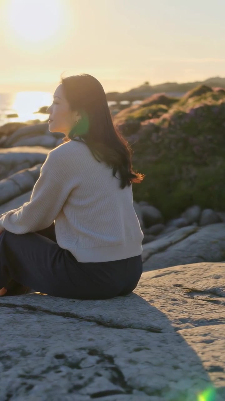 Video verticale: Donna seduta che guarda il sole tramontare dalla riva con erica, indossando un maglione beige.