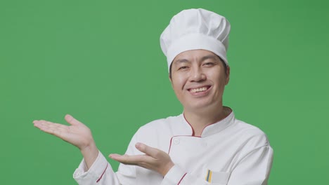primer plano de un chef asiático sonriendo y señalando hacia un lado mientras está de pie en el estudio de fondo de la pantalla verde