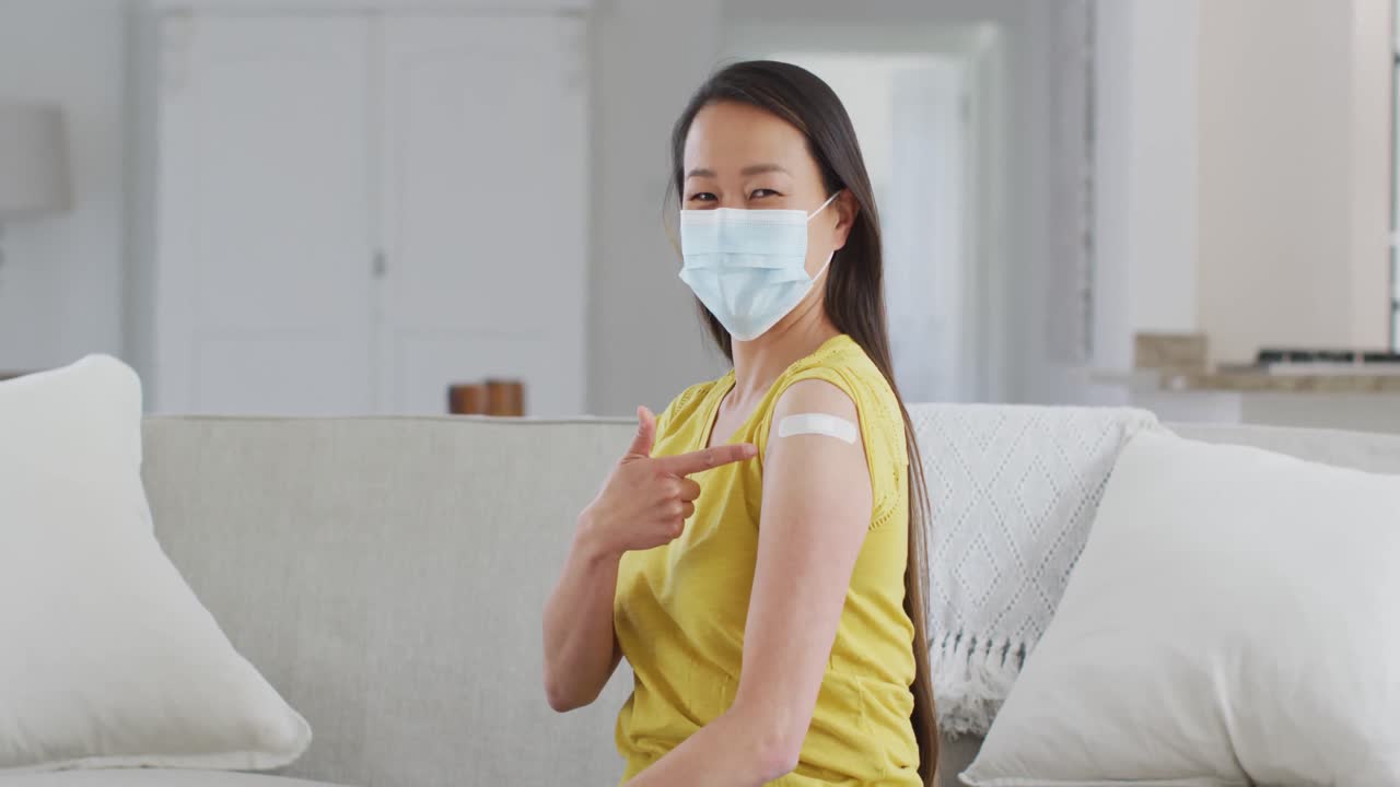 Happy asian woman in face mask sitting on sofa showing arm with plaster after vaccination