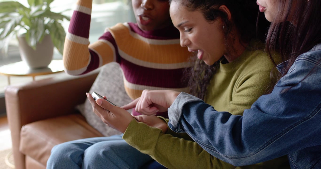 Diverse female pals leaning as mid woman extending arm holding phone while checking selfies on sofa
