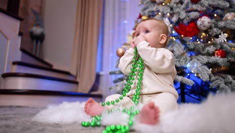 cute little child wearing white clothes sits on the floor near the christmas tree. adorable baby plays with beads decorations for the holidays. low angle view.