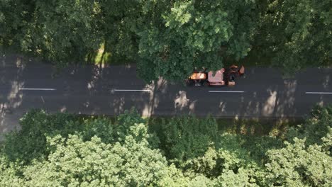 a top-down aerial view shows a maintenance vehicle partially hidden by tree canopies while moved on a narrow forest road. a black car passes by on the adjacent lane.