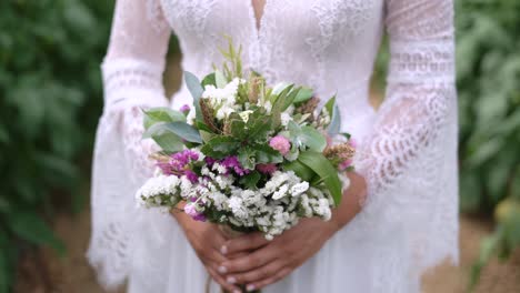 Beautiful-Bride-holding-wildflowers-bouquet-in-hand-outdoor-lush-foliage-Background