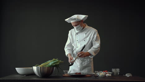 chef masculino con máscara facial médica, usando un cepillo de silicona con salsa en un plato de carne gourmet. cocinero profesional trabajando en la cocina.