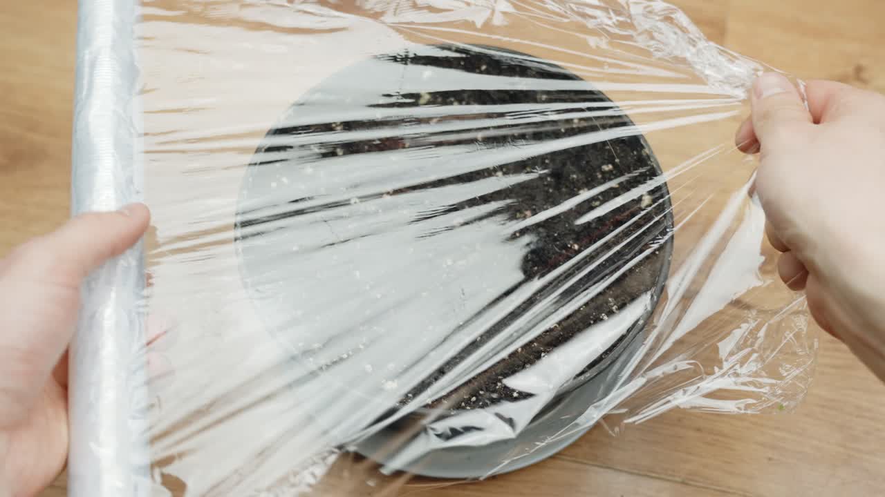 Person covering flowerpot full of dark soil and seed with plastic to grow