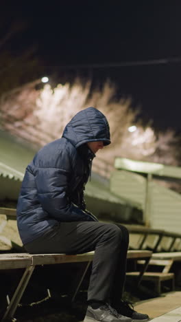 후드를 입은 남자가 혼자 경기장 관중석에 앉아 있으며, 흐릿하고 희미하게 조명된 배경이 있습니다.