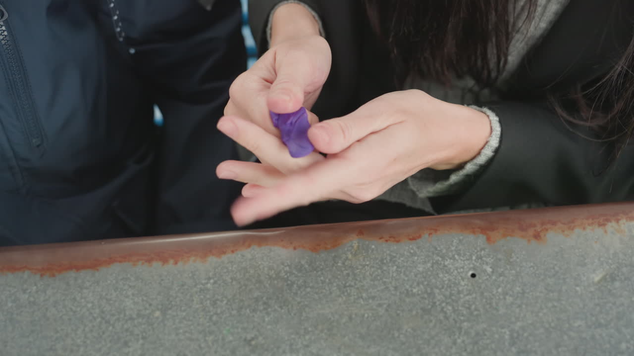 Student hands shaping purple clay together, classroom desk edge and colorful clay kit visible, teacher guiding small hands through tactile modeling, preparing backtoschool art project, closeup