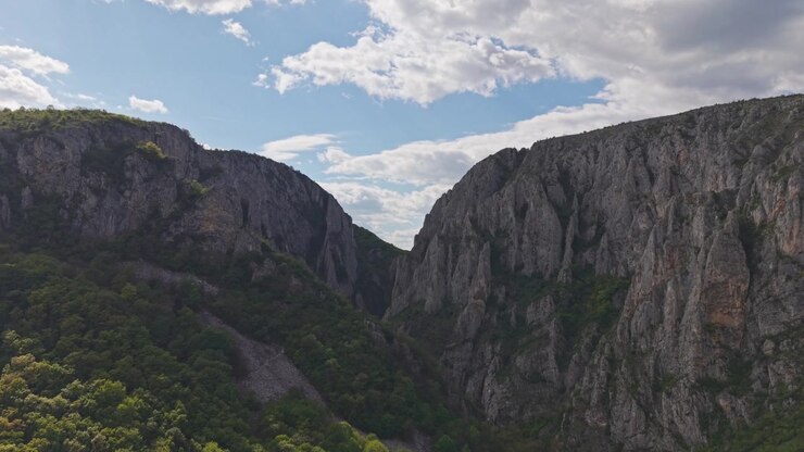 Vista panorâmica de um cânion de montanha dramático cercado por densa floresta verde sob um céu azul brilhante com nuvens