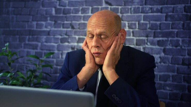 A bored business man with head in hands staring into space pensive in front of a computer laptop in an office at work