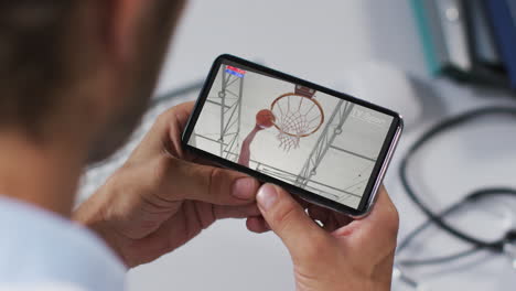 compuesto de un médico masculino viendo un juego de baloncesto en un teléfono inteligente