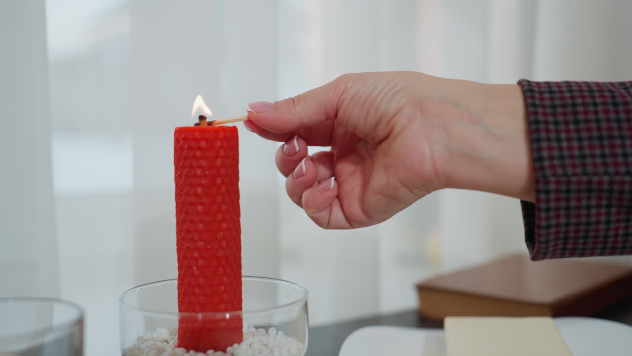 primer plano de la mano encendiendo una vela roja con cerilla en la mesa de la cocina, el brillo cálido agrega un ambiente acogedor, con mantequilla, platos y vidrios en el fondo