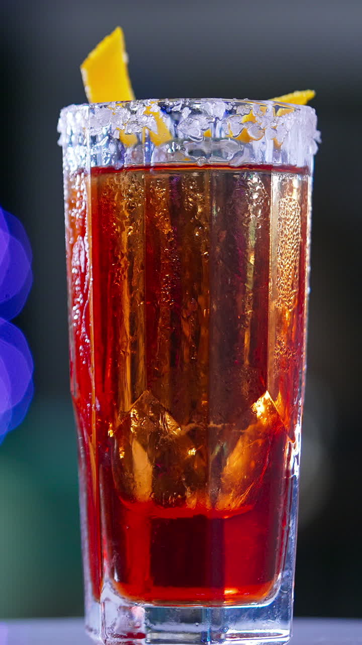 Misted long glass with red drink. Rotating table with cocktail decorated with orange peel. Close up. Blurred backdrop. Vertical video