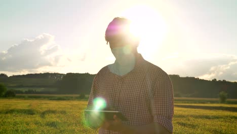 joven agricultor confiado en la camisa sosteniendo la tableta y mirando hacia adelante en el campo de trigo, hermosa tierra cultivada en el fondo