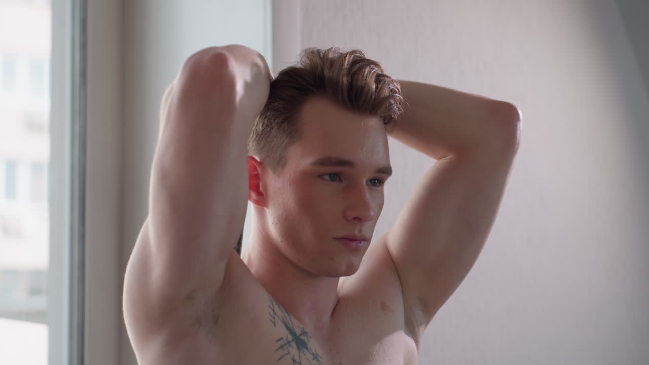 focused gym lover stretches resistance band overhead while standing beside window, sunlight casting soft highlights across chest and arms, showcasing tattoo and toned physique in quiet indoor setting