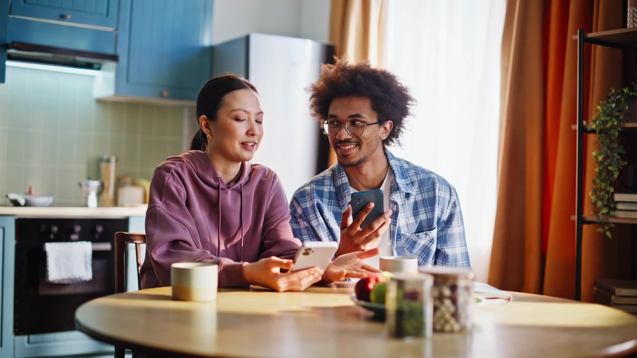 Couple sharing breakfast in kitchen. Mixed race pair browsing smartphones