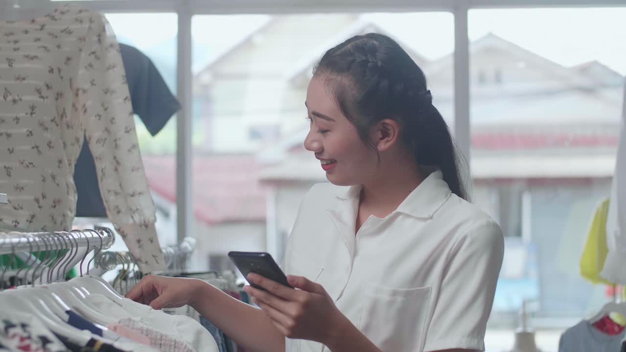 부티크의 커에 있는 다양한 의류를 선택하는 동안 스마트폰을 사용하는 미소 짓는 젊은 아시아 여성