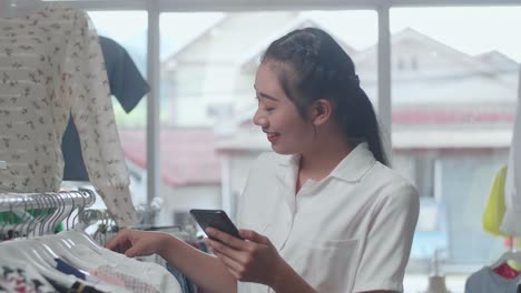 smiling young asian woman using smartphone while choosing various clothing items on hangers at the boutique