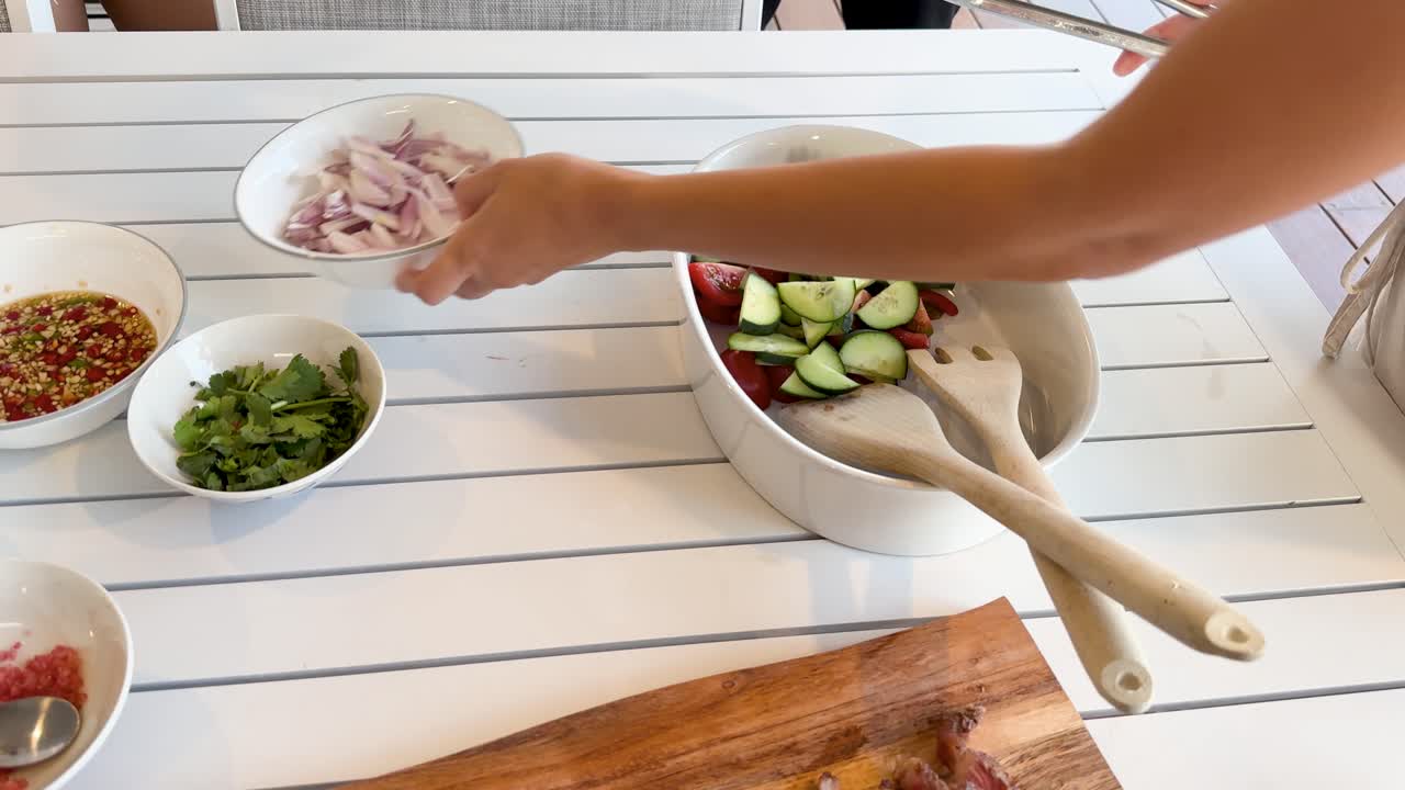 A person adds sliced vegetables and herbs to a salad bowl on a bright, sunlit table, assembling a Thai beef salad with fresh ingredients
