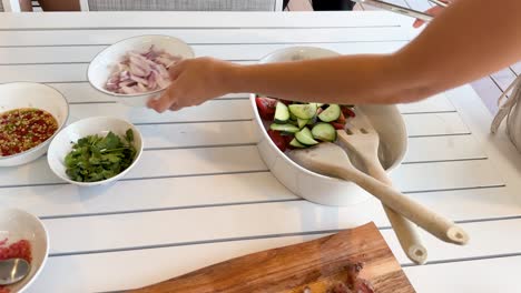 a person adds sliced vegetables and herbs to a salad bowl on a bright, sunlit table, assembling a thai beef salad with fresh ingredients