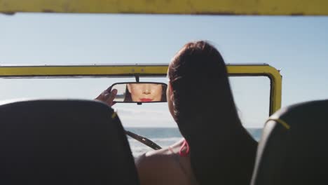 Happy-caucasian-woman-sitting-in-beach-buggy-by-the-sea-doing-make-up