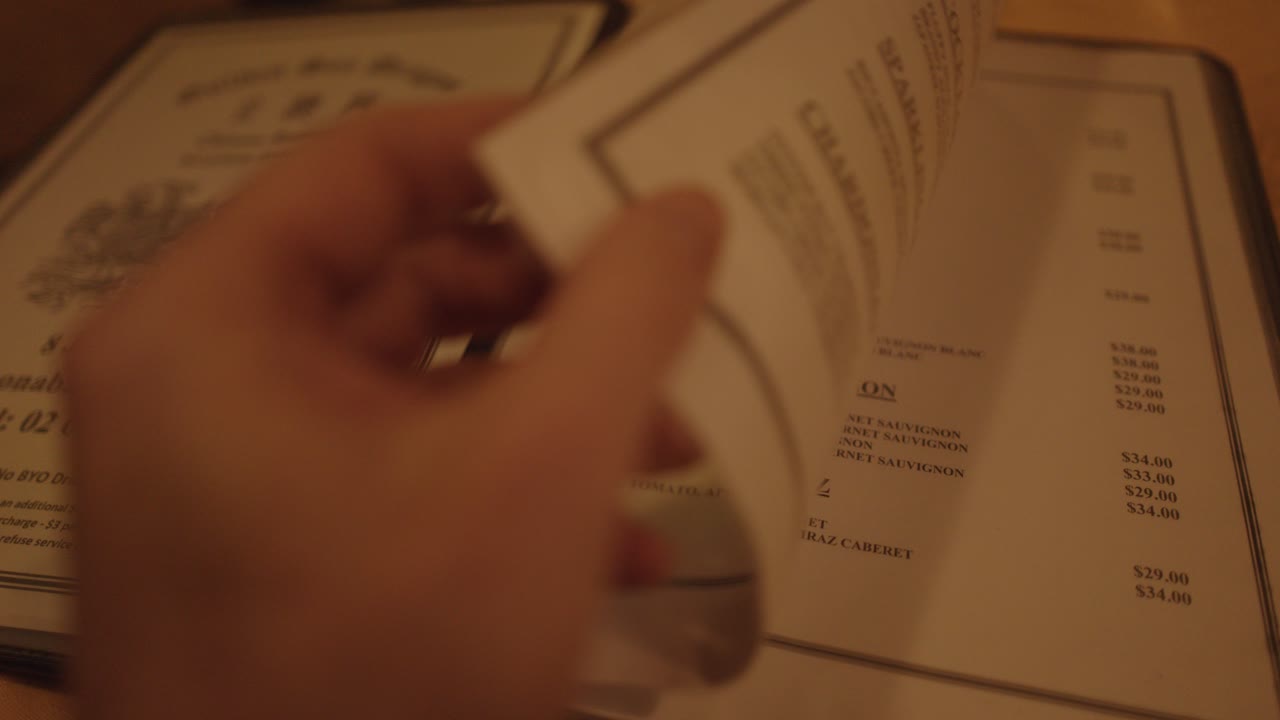 A person turns pages of a restaurant menu under soft, ambient lighting at a table