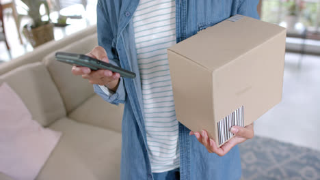 midsection of biracial woman holding parcel and using smartphone at home, slow motion