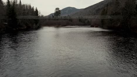 Flying-Over-Calm-Lake-Under-A-Bridge-With-Tourist-Crossing-During-Winter-In-Vallee-Bras-du-Nord-At-Saint-Raymond,-Canada