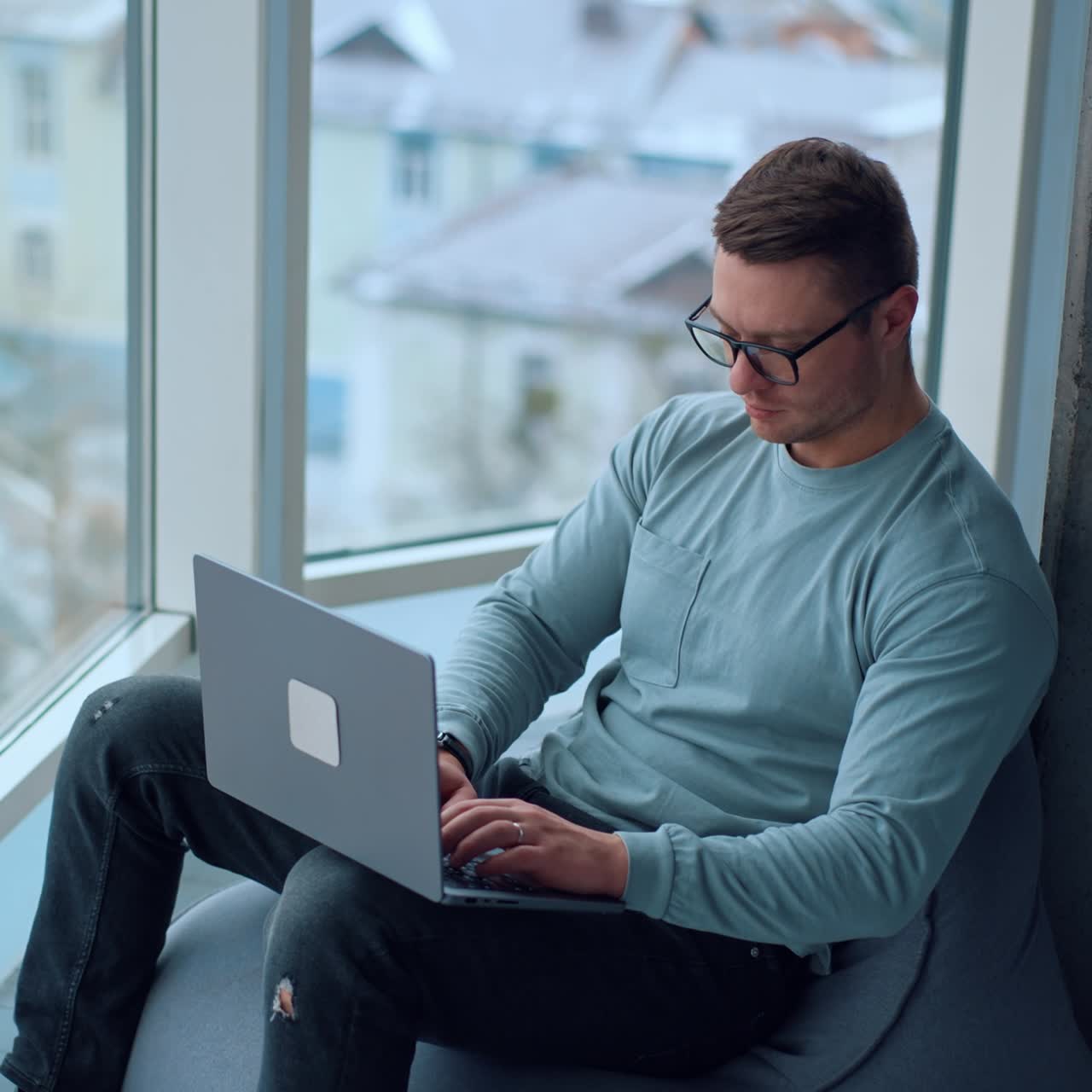 Freelancing young adult man concentrated on the work on laptop. Man in glasses sits near the window working on computer. View from top