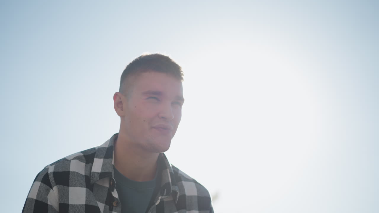 sunlight creates silhouette glow around fair skinned man in checkered shirt as he lifts hand to mouth while gently closing eyes in bright outdoor setting under soft sky during calm moment