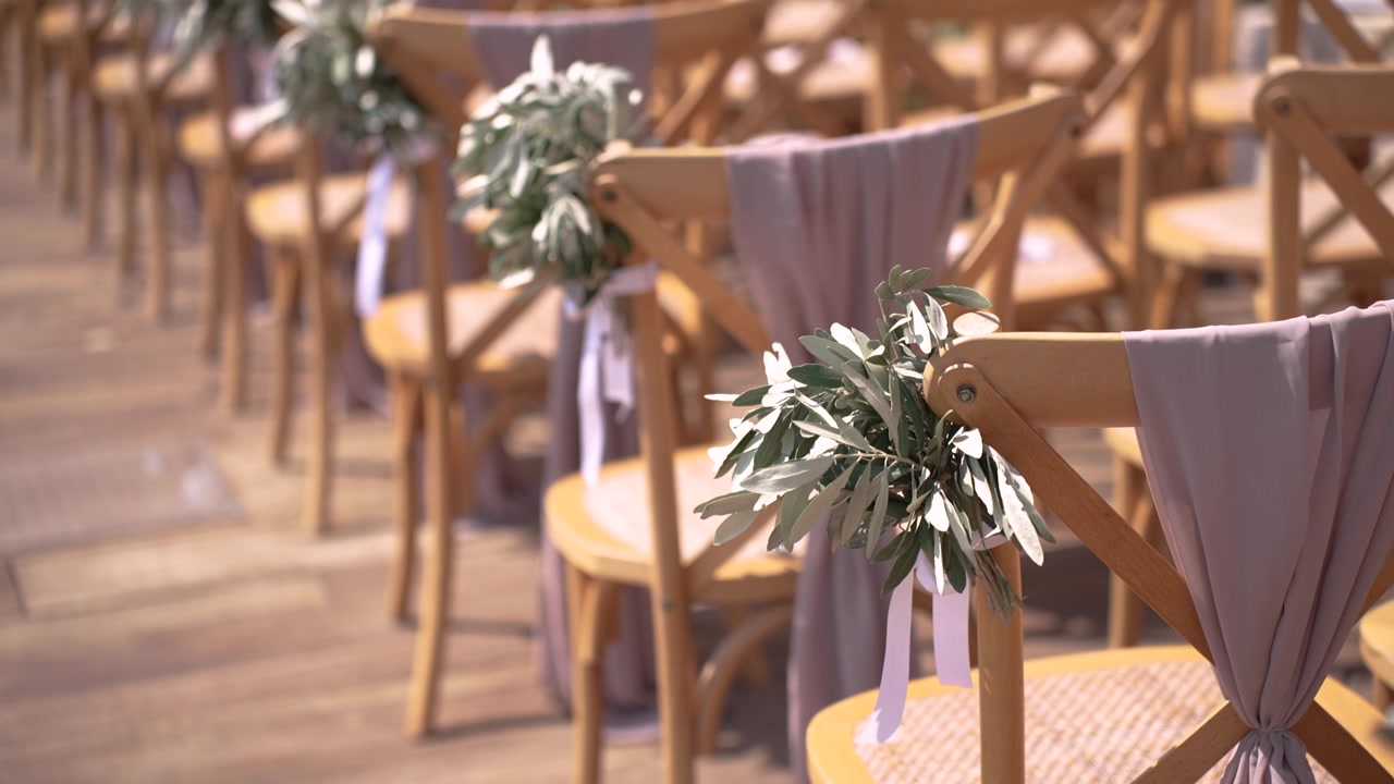 Le sedie sono disposte in file per una cerimonia di matrimonio. Ogni sedia ha del verde legato con un nastro sullo schienale. L'allestimento è pronto per gli ospiti mentre si avvicina l'ora dell'evento.