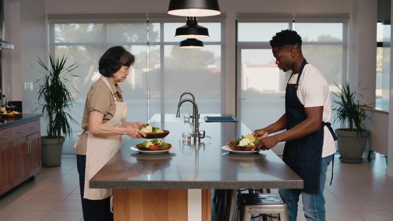 Passing woman extending salad bowl to man, plating salads on kitchen island with aprons