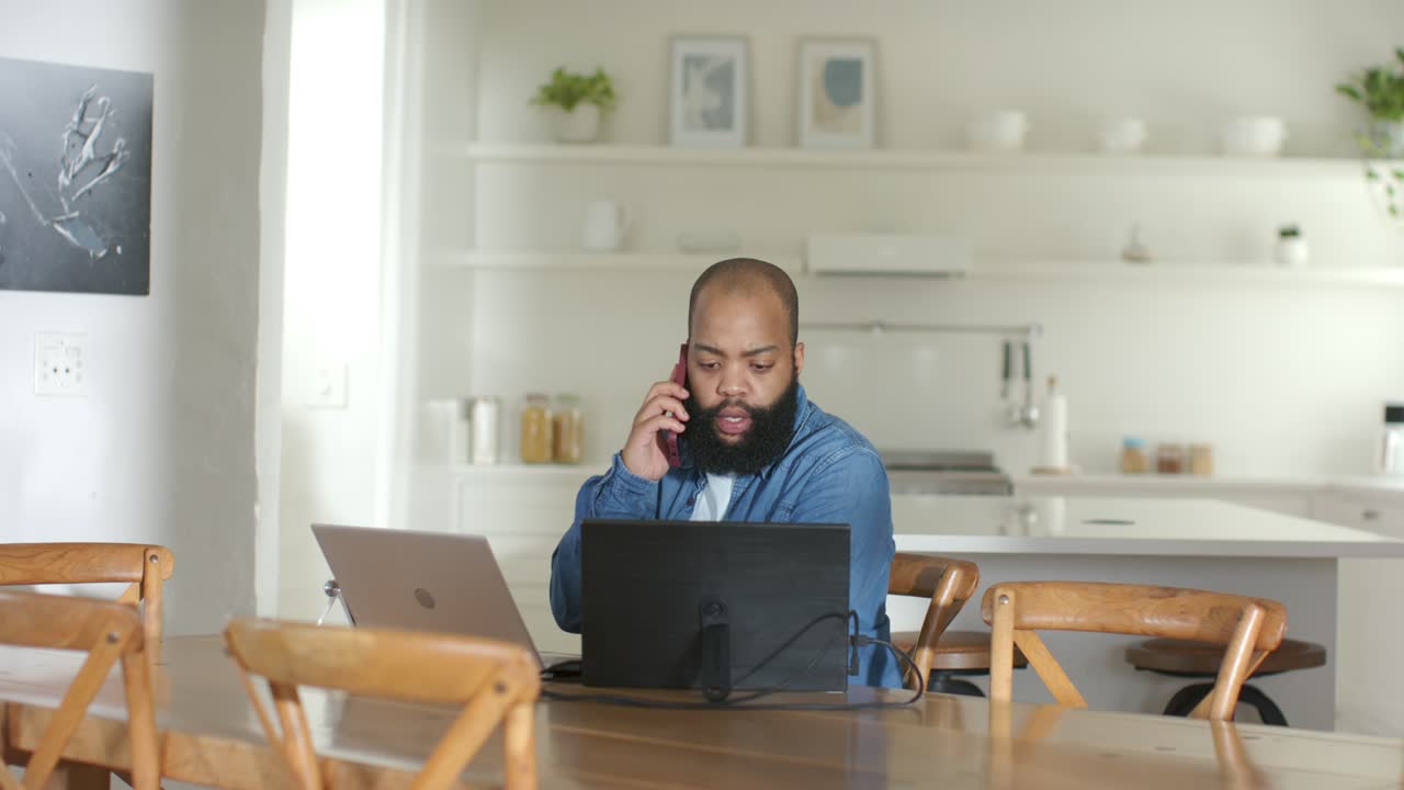 Vertical video: Taking work call, man in denim toggling between phone and laptop at dining table