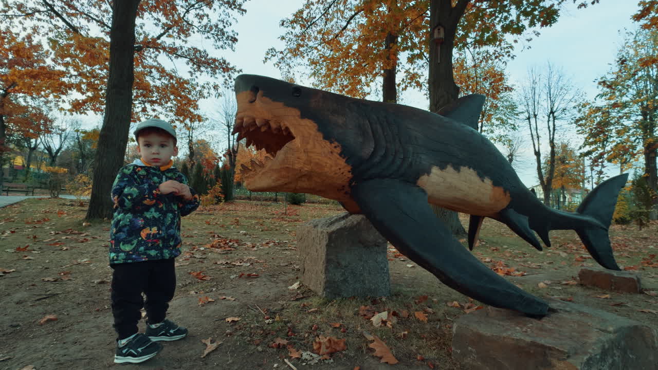 Caucasian toddler playing at the life-size wooden sculpture of a shark. in the park. Little boy puts something into the jaws and turns to camera.