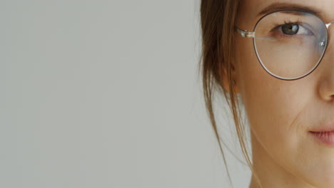 Close-Up-Of-The-Half-Female-Facen-In-Glasses-With-Long-Fair-Hair-Looking-In-The-Camera-And-Then-Smiling-On-The-White-Wall-Background