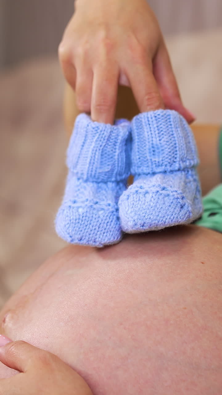Pregnant woman holding baby booties at home, closeup. Eight months pregnant woman with blue booties on her belly. Vertical video