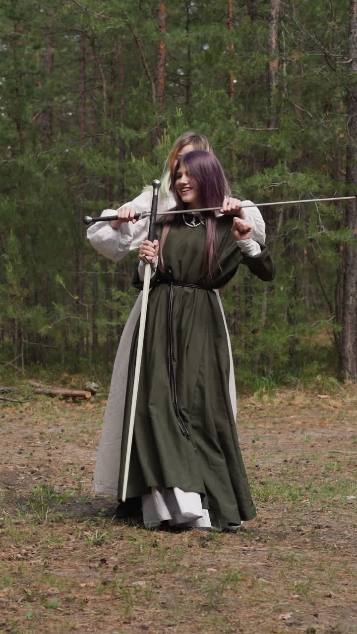 una adolescente con un vestido medieval baila mientras su amiga sostiene la espada por el cuello en el bosque de abeto en cámara lenta. juego de rol para niños. reconstrucción de armas antiguas