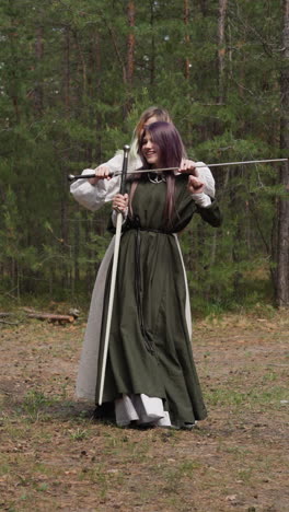 girl teenager in medieval dress dances while friend holds sword by neck in fir forest slow motion. role playing for children. old weapon reconstruction