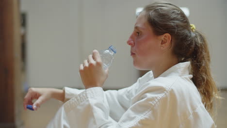 vista lateral de una chica exhausta con kimono bebiendo agua en el gimnasio