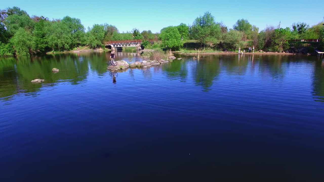 Beautiful natural view on bright sunny summer day. Drone flying over the calm blue river with green wooded waterfront. People spending time in the nature outside the city.