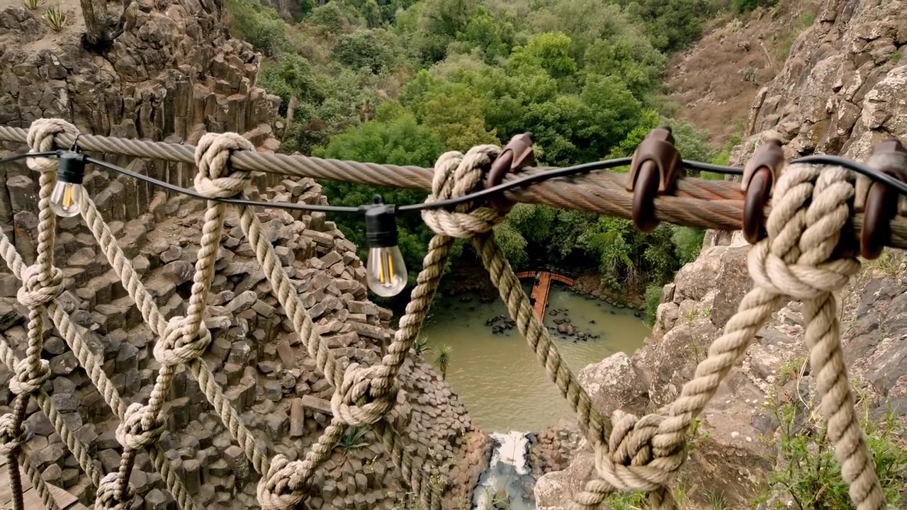 High angle view looking down into basalt rock canyon featuring small bridge, water and dense green trees in natural landscape