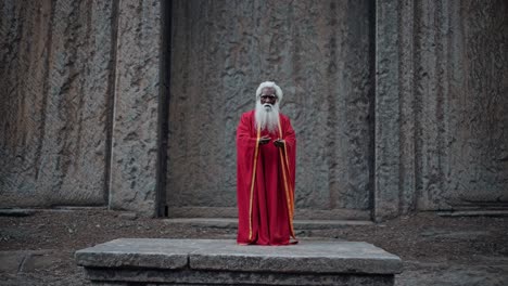 senior orthodox priest wearing ornate red gold vestments, standing with clasped hands near rock carved lalibela church, historic ethiopian christian landmark designated unesco world heritage site