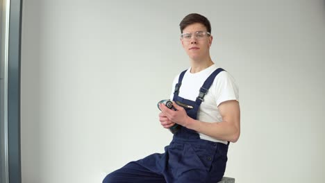 young craftsman in work uniform and with a screwdriver in his hands sits on a white wall background